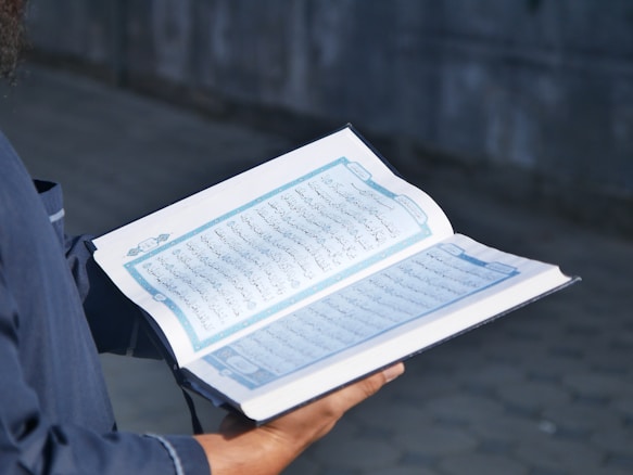 A person holding an open book with Arabic script on its pages. The book appears to be religious in nature, possibly the Quran. The person is wearing a dark blue garment with visible sleeves.