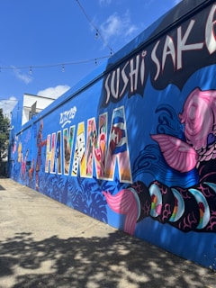 A vibrant mural on a wall in an urban setting features the words 'Little Havana' in large, colorful letters filled with a variety of patterns. The mural includes intricate designs of fish, leaves, and other artistic elements. Overhead, there are string lights, and the sky is clear with a few clouds.