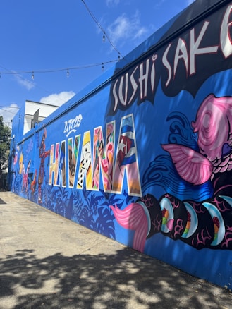 A vibrant mural on a wall in an urban setting features the words 'Little Havana' in large, colorful letters filled with a variety of patterns. The mural includes intricate designs of fish, leaves, and other artistic elements. Overhead, there are string lights, and the sky is clear with a few clouds.