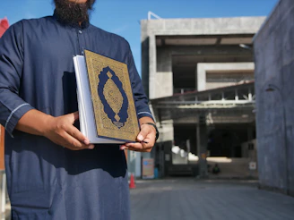A person wearing a dark blue traditional robe is holding an ornate book with a blue and gold cover featuring intricate designs. The background shows a modern concrete building, partially shaded.