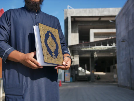 A person wearing a dark blue traditional robe is holding an ornate book with a blue and gold cover featuring intricate designs. The background shows a modern concrete building, partially shaded.