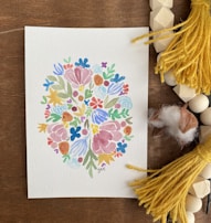 A watercolor painting features an intricate pattern of colorful flowers and leaves arranged in a symmetrical, abstract design. The artwork is placed on a wooden surface surrounded by yellow tassels, wooden beads, and pieces of soft cotton.