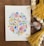 A watercolor painting features an intricate pattern of colorful flowers and leaves arranged in a symmetrical, abstract design. The artwork is placed on a wooden surface surrounded by yellow tassels, wooden beads, and pieces of soft cotton.