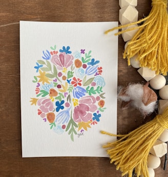 A watercolor painting features an intricate pattern of colorful flowers and leaves arranged in a symmetrical, abstract design. The artwork is placed on a wooden surface surrounded by yellow tassels, wooden beads, and pieces of soft cotton.