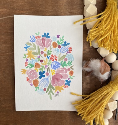 A watercolor painting features an intricate pattern of colorful flowers and leaves arranged in a symmetrical, abstract design. The artwork is placed on a wooden surface surrounded by yellow tassels, wooden beads, and pieces of soft cotton.