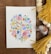 A watercolor painting features an intricate pattern of colorful flowers and leaves arranged in a symmetrical, abstract design. The artwork is placed on a wooden surface surrounded by yellow tassels, wooden beads, and pieces of soft cotton.