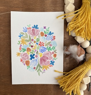 A watercolor painting features an intricate pattern of colorful flowers and leaves arranged in a symmetrical, abstract design. The artwork is placed on a wooden surface surrounded by yellow tassels, wooden beads, and pieces of soft cotton.