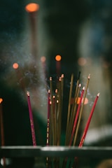 A group of lit incense sticks with smoke gently rising in a dimly lit setting. The focus is soft, highlighting the warm glow of the incense tips against a dark and blurred background, creating a serene and spiritual ambiance.
