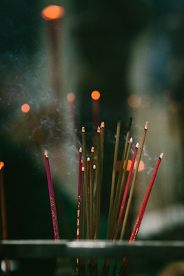 A group of lit incense sticks with smoke gently rising in a dimly lit setting. The focus is soft, highlighting the warm glow of the incense tips against a dark and blurred background, creating a serene and spiritual ambiance.