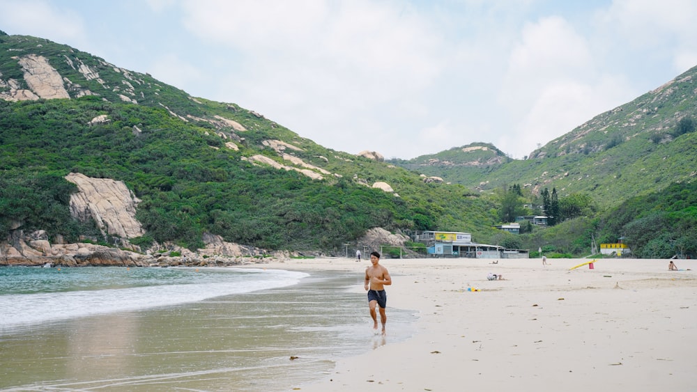 a man walking along a beach next to a lush green hillside