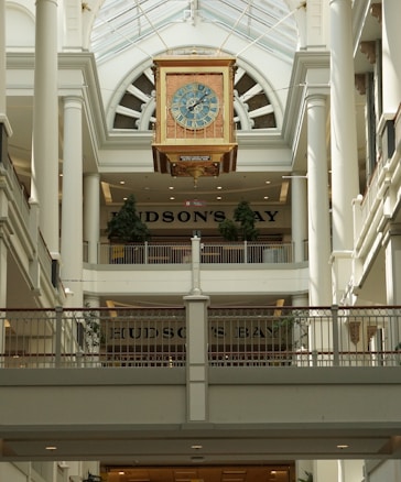 An ornate clock with a detailed face is suspended in the center of an elegant shopping mall atrium. The clock is surrounded by tall white columns and railings, with the store name visible in the background. Natural light streams through a large glass roof.