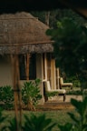 A cozy cottage with a thatched roof surrounded by lush greenery. Wooden chairs are placed on the patio, adding to the serene and rustic charm. The foliage around the house gives a natural and secluded feel.