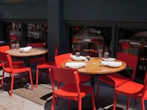 Several wooden tables with red chairs are set up outdoors. Each table is neatly arranged with white napkins, plates, and clear drinking glasses. The tables are positioned on a pavement, next to a dark green building with windows reflecting the street scenery.