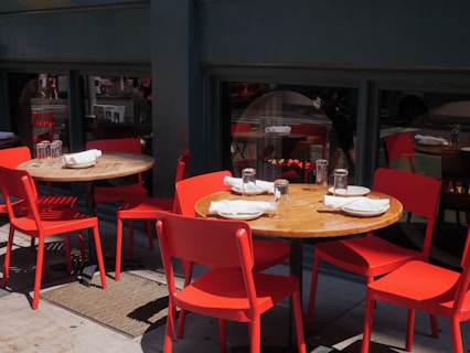 Several wooden tables with red chairs are set up outdoors. Each table is neatly arranged with white napkins, plates, and clear drinking glasses. The tables are positioned on a pavement, next to a dark green building with windows reflecting the street scenery.