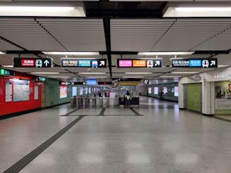 An underground subway station with multiple signs indicating various train lines. The station is clean and well-lit with overhead lights and a tiled floor. There are directional signs with colorful labels above a set of automated ticket gates. The walls display maps and advertisements. A few people are visible near the ticket gates.