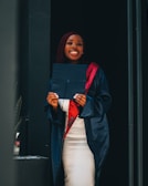 A person wearing a graduation gown and cap holds a diploma while smiling broadly. The gown is a deep shade of navy blue with a red stole. The background is simple and dark, highlighting the person.