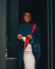 A person wearing a graduation gown and cap holds a diploma while smiling broadly. The gown is a deep shade of navy blue with a red stole. The background is simple and dark, highlighting the person.