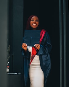 A person wearing a graduation gown and cap holds a diploma while smiling broadly. The gown is a deep shade of navy blue with a red stole. The background is simple and dark, highlighting the person.