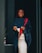 A person wearing a graduation gown and cap holds a diploma while smiling broadly. The gown is a deep shade of navy blue with a red stole. The background is simple and dark, highlighting the person.