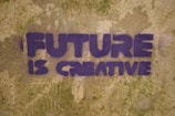 Purple graffiti text on a textured, weathered wall reads 'FUTURE IS CREATIVE'. The letters are bold and blocky, set against a mottled tan and green background.