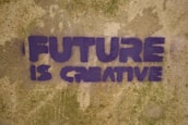 Purple graffiti text on a textured, weathered wall reads 'FUTURE IS CREATIVE'. The letters are bold and blocky, set against a mottled tan and green background.