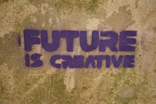 Purple graffiti text on a textured, weathered wall reads 'FUTURE IS CREATIVE'. The letters are bold and blocky, set against a mottled tan and green background.