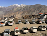 A scenic mountain landscape with several parked vehicles, including buses and SUVs, on a dirt lot. The rocky cliffs and rugged peaks are seen in the background, with buildings having orange rooftops nearby.