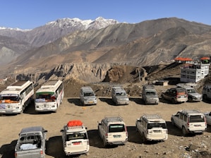 A scenic mountain landscape with several parked vehicles, including buses and SUVs, on a dirt lot. The rocky cliffs and rugged peaks are seen in the background, with buildings having orange rooftops nearby.