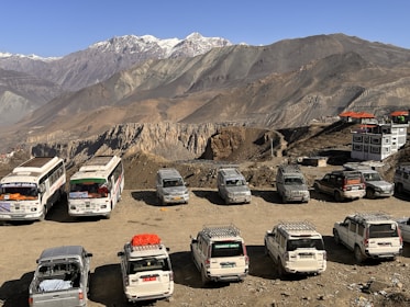 A scenic mountain landscape with several parked vehicles, including buses and SUVs, on a dirt lot. The rocky cliffs and rugged peaks are seen in the background, with buildings having orange rooftops nearby.