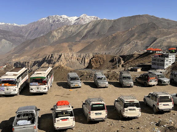 A scenic mountain landscape with several parked vehicles, including buses and SUVs, on a dirt lot. The rocky cliffs and rugged peaks are seen in the background, with buildings having orange rooftops nearby.
