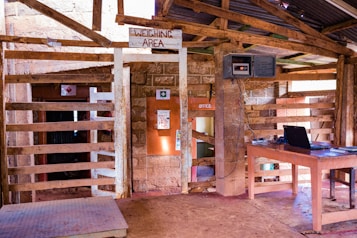 An interior setting with wooden panel structures, a table with a laptop, a wall-mounted digital display, and a sign reading 'Weighing Area'. Sunlight illuminates the rustic wooden and stone materials. A small opening labeled 'Office' is visible, alongside a first aid sign.