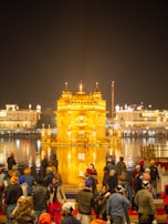 A large group of people gathers near a beautifully illuminated golden temple, reflected in a surrounding body of water. The temple's intricate architecture is highlighted by bright lighting, and the night sky provides a dark backdrop.