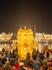 A large group of people gathers near a beautifully illuminated golden temple, reflected in a surrounding body of water. The temple's intricate architecture is highlighted by bright lighting, and the night sky provides a dark backdrop.