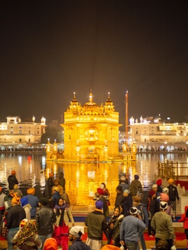 A large group of people gathers near a beautifully illuminated golden temple, reflected in a surrounding body of water. The temple's intricate architecture is highlighted by bright lighting, and the night sky provides a dark backdrop.