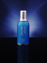 A blue bottle with a silver cap is centered against a dark blue gradient background. The bottle features white text displaying its brand and product name.