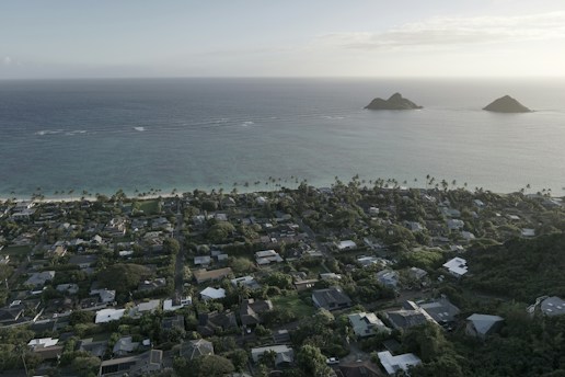 A coastal residential area with numerous houses surrounded by lush greenery and palm trees, leading up to a sandy beach along a calm ocean. Two small islands are visible in the distance, under a clear sky.
