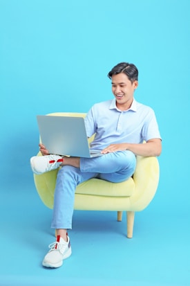 A person sitting on a yellow armchair, casually looking at a laptop. They are smiling, wearing a light blue shirt and matching pants, with white sneakers. The background is a solid blue color that complements the overall theme.