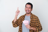 A smiling man holds a fan of banknotes in one hand, while pointing upwards with the other. He wears a yellow and black plaid shirt over a gray t-shirt, against a neutral background.