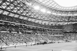 A large stadium filled with spectators sits under a vast, intricately designed roof. Bright lights illuminate the area, highlighting thousands of people seated in multiple tiers. The architecture features a web-like pattern supporting the ceiling, creating an open and expansive atmosphere.