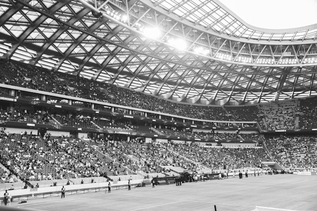 A large stadium filled with spectators sits under a vast, intricately designed roof. Bright lights illuminate the area, highlighting thousands of people seated in multiple tiers. The architecture features a web-like pattern supporting the ceiling, creating an open and expansive atmosphere.
