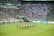 A large sports stadium is filled with spectators, the majority wearing green shirts, creating a vibrant and energetic atmosphere. In the foreground, a group of individuals dressed in blue shirts are carrying a large flag across the field. Stadium staff and officials are positioned near the field and among the crowd.
