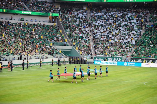 A large sports stadium is filled with spectators, the majority wearing green shirts, creating a vibrant and energetic atmosphere. In the foreground, a group of individuals dressed in blue shirts are carrying a large flag across the field. Stadium staff and officials are positioned near the field and among the crowd.