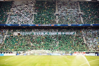 A large crowd fills a sports stadium, with many people wearing green shirts. The fans are densely packed, with some holding banners displaying text. The stadium seating is visible, and the field has sprinklers turned on, soaking the grass.