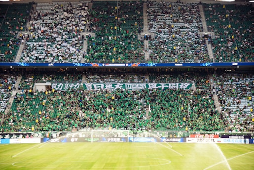 A large crowd fills a sports stadium, with many people wearing green shirts. The fans are densely packed, with some holding banners displaying text. The stadium seating is visible, and the field has sprinklers turned on, soaking the grass.