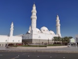 The image features a large, white mosque with multiple domes and four tall minarets, set against a clear blue sky. The architecture is ornate, with detailed carvings and a pristine facade. A fenced garden area with palm trees surrounds the building, and a few people can be seen walking nearby.