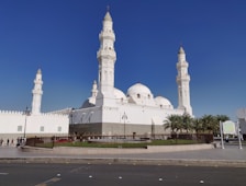 The image features a large, white mosque with multiple domes and four tall minarets, set against a clear blue sky. The architecture is ornate, with detailed carvings and a pristine facade. A fenced garden area with palm trees surrounds the building, and a few people can be seen walking nearby.
