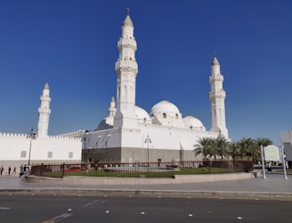 The image features a large, white mosque with multiple domes and four tall minarets, set against a clear blue sky. The architecture is ornate, with detailed carvings and a pristine facade. A fenced garden area with palm trees surrounds the building, and a few people can be seen walking nearby.