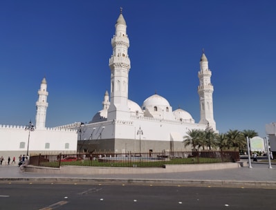 The image features a large, white mosque with multiple domes and four tall minarets, set against a clear blue sky. The architecture is ornate, with detailed carvings and a pristine facade. A fenced garden area with palm trees surrounds the building, and a few people can be seen walking nearby.