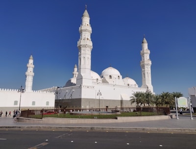 The image features a large, white mosque with multiple domes and four tall minarets, set against a clear blue sky. The architecture is ornate, with detailed carvings and a pristine facade. A fenced garden area with palm trees surrounds the building, and a few people can be seen walking nearby.