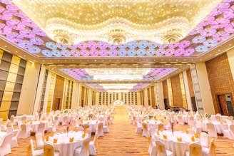 A large, elegantly decorated banquet hall with numerous round tables covered with white tablecloths and adorned with flowers. The ceiling features intricate lighting with patterns in shades of purple and gold. The floor appears to be a glossy wood finish under soft lighting, giving the room a luxurious feel.